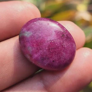 Puede incluir: Una piedra preciosa pulida, de forma ovalada, en tonos magenta oscuro y rojo violáceo. La piedra tiene una superficie lisa y brillante con sutiles variaciones de color y textura. La piedra preciosa se sostiene en una mano, con un fondo borroso verde y amarillo.