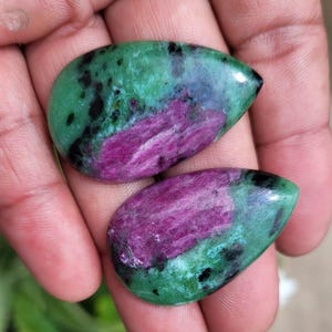 Może przedstawiać: Dwa polerowane kamienie szlachetne z rubinu zoisytu. Każdy kamień ma kształt łzy, z żywym zielonym kolorem bazowym nakrapianym czernią i centralną smugą głębokiej magenty. Kamienie są trzymane w dłoni.