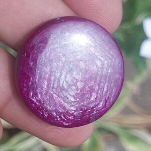 Puede incluir: Una gema redonda y pulida con un tono magenta intenso, que exhibe una superficie texturizada y un sutil juego de luces. El color de la gema transita de un magenta vibrante en los bordes a un centro más claro, casi blanco.