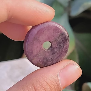 以下が含まれることがあります： 紫とピンクの色合いの、丸く磨かれた宝石ドーナツが指の間に挟まれています。 石の中心には穴があります。 背景はぼやけた緑の葉です。