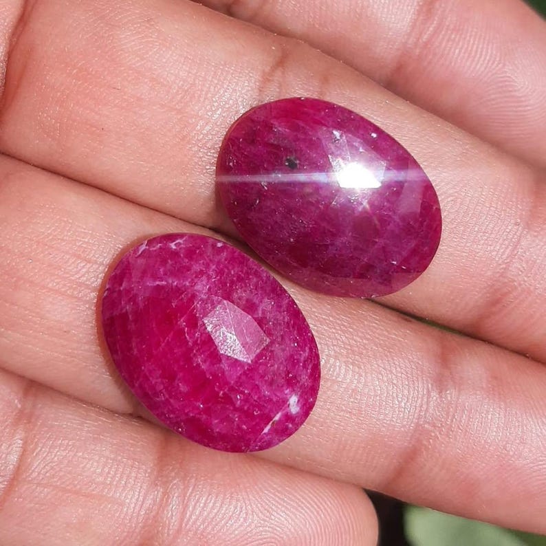 May include: Two oval-shaped, polished ruby gemstones. One ruby has a star effect, with a white line across the center. The other ruby has a faceted surface. Both gems are a deep, rich red color.