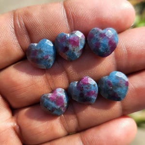 Könnte beinhalten: Sechs herzförmige Edelsteine mit facettierter Oberfläche. Die Steine sind eine Mischung aus Blau und Magenta. Die Edelsteine werden in einer Hand gehalten.