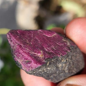 以下が含まれることがあります： 未研磨の原石の破片。片面は鮮やかで深いマゼンタピンクの結晶面になっており、不規則な形状の石の残りの部分は、暗く粗い質感のチャコールグレーの鉱物マトリックスで構成されています。