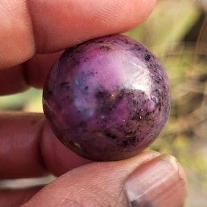 Peut inclure: Une pierre précieuse sphérique polie, d'un magenta profond, parsemée d'inclusions gris foncé et noires. La pierre est tenue entre deux doigts, mettant en valeur sa surface lisse et réfléchissante et ses variations naturelles de couleur.