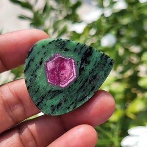 May include: A teardrop-shaped zoisite gemstone featuring a vibrant, hexagonal magenta ruby crystal embedded in the centre. The stone displays a mottled green base with dark black mineral inclusions throughout the surface texture.