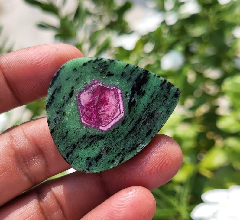 May include: A teardrop-shaped zoisite gemstone featuring a vibrant, hexagonal magenta ruby crystal embedded in the centre. The stone displays a mottled green base with dark black mineral inclusions throughout the surface texture.