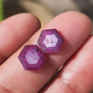 May include: Two hexagonal-cut, deep magenta gemstones with a lighter center are displayed. The gems have a faceted design and dark inclusions. The gems are held in a hand, showcasing their color and shape. These gems are suitable for jewelry making.