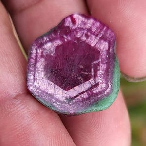 May include: A close-up of a gemstone with a hexagonal centre. The gem features a deep magenta hue in the centre, transitioning to lighter shades towards the edges. A thin green border surrounds the gem.
