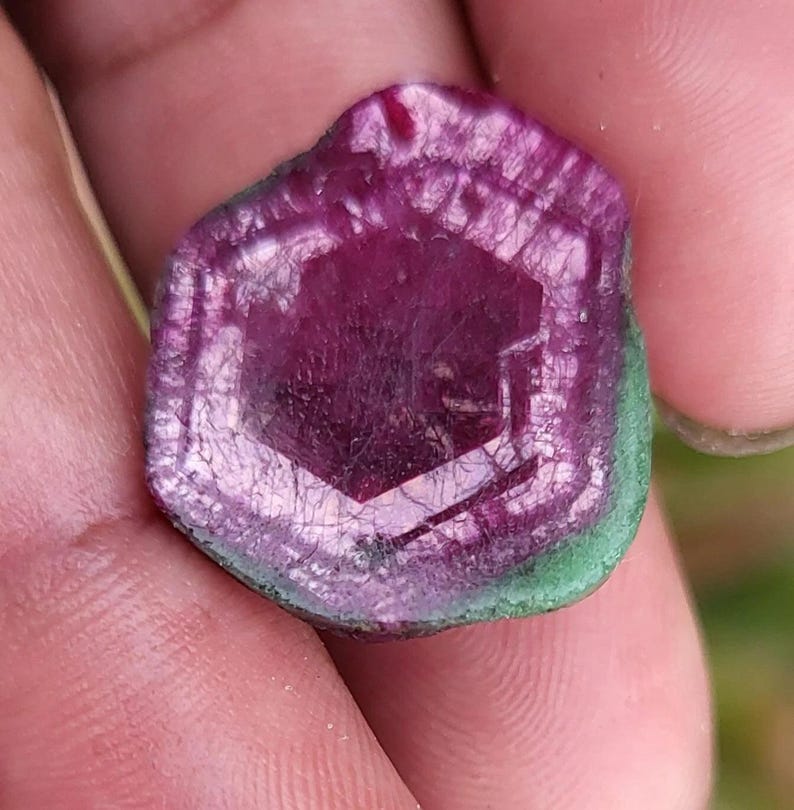 May include: A close-up of a gemstone with a hexagonal centre. The gem features a deep magenta hue in the centre, transitioning to lighter shades towards the edges. A thin green border surrounds the gem.