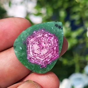 以下が含まれることがあります： 鮮やかなマゼンタ色の六角形の芯を持ち、周囲を緑色の縁で囲まれた、涙型の研磨された宝石。中心部には、ピンクと濃い赤の色合いで層状になった幾何学模様の独特な結晶構造が見られます。