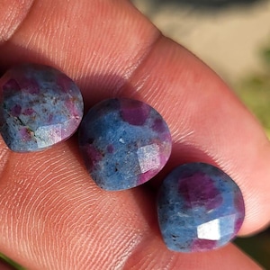 May include: Three faceted gemstones with a mix of blue and red hues. The stones have a rounded shape and are held in a hand. The gems have a polished, reflective surface.