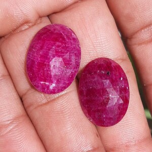 May include: Two faceted, oval-shaped rubies. The gemstones are a deep, vibrant red with a slight pink hue. The cut of the rubies creates multiple facets that reflect light, enhancing their brilliance. The rubies are set against a neutral background.