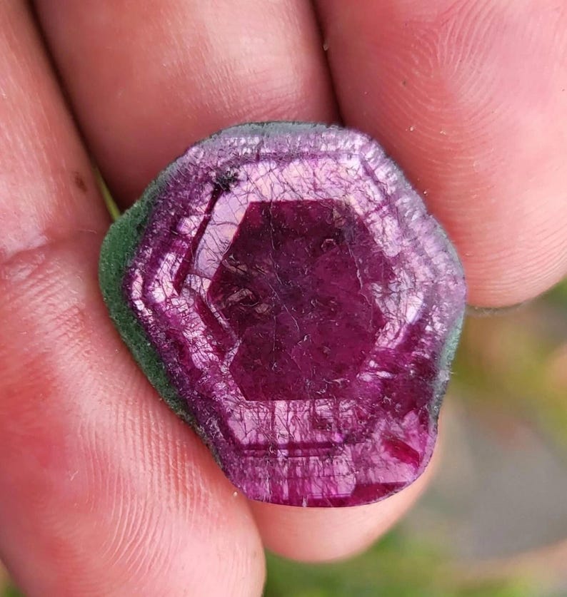 May include: A close-up of a gemstone with a hexagonal shape and a deep magenta hue. The gem has a green outer edge and a crystalline structure, showcasing natural textures and patterns. The gem is held in a hand.