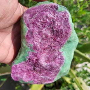 Könnte beinhalten: Ein grob geschnittener Edelstein mit einem leuchtend magenta Zentrum, das von einem grünen Äußeren umgeben ist. Die Oberfläche des Steins hat ein strukturiertes Aussehen mit sichtbaren kristallinen Strukturen. Der Naturstein wird in einer Hand gehalten.