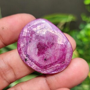 May include: A polished, oval gemstone in a deep magenta colour. The stone has a faceted surface that reflects light, creating a sparkling effect. Small black inclusions are visible. The gemstone is held in a hand.