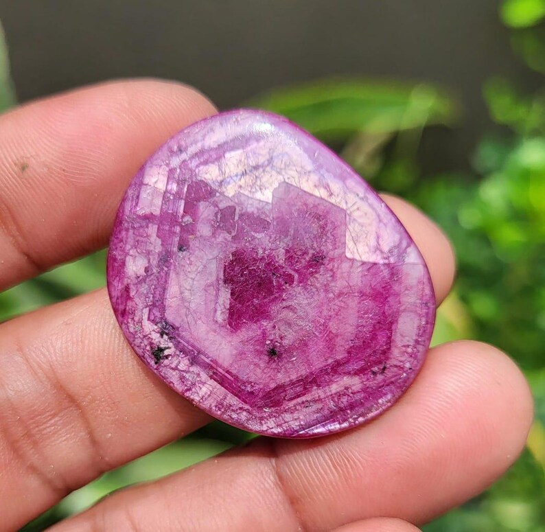 May include: A polished, oval gemstone in a deep magenta colour. The stone has a faceted surface that reflects light, creating a sparkling effect. Small black inclusions are visible. The gemstone is held in a hand.