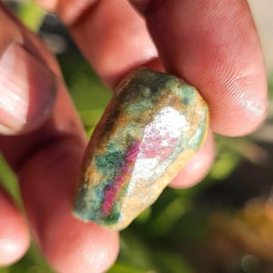 May include: A natural gemstone with a mix of colours, including green, red, and tan. The stone is held in a hand, and the surface of the stone is polished and reflective. The gemstone is likely a type of mineral specimen.