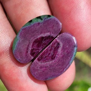 K&ouml;nnte beinhalten: Zwei polierte Rubin-Zoisit-Edelsteine, oval geformt, mit tiefmagentafarbenem Inneren und gr&uuml;nem Rand. Die Edelsteine sind gespalten und zeigen die inneren Muster und Texturen. Die Edelsteine werden in einer Hand gehalten.
