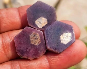 Puede incluir: Tres gemas hexagonales en bruto en tonos morados y violetas. Cada piedra tiene una inclusión más clara, de seis lados, en el centro. Las gemas se sostienen en una mano, con un fondo borroso de color verde.