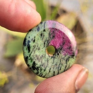 Può includere: Un ciondolo rotondo in pietra lucida con un foro centrale. La pietra presenta un mix di colori verde, nero e magenta. Il ciondolo è tenuto tra due dita.