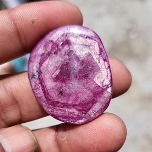 May include: A polished, oval gemstone in a deep magenta colour, displaying intricate internal patterns and a slightly translucent appearance. The stone is held between fingers, highlighting its natural beauty and unique veining.