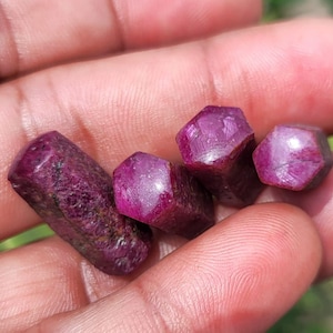 Puede incluir: Cuatro cristales de rubí en bruto y sin pulir con un tono rojo magenta intenso. Las piedras presentan formas prismáticas hexagonales naturales con texturas variadas, que van desde superficies rugosas y opacas hasta facetas cristalinas lisas.