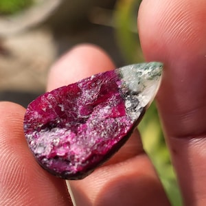 Peut inclure: Une pierre précieuse brute, d'un rouge profond, avec une surface texturée, virant à une teinte plus claire et translucide à l'extrémité. La pierre est tenue dans une main. La forme est irrégulière, avec un mélange de couleurs.