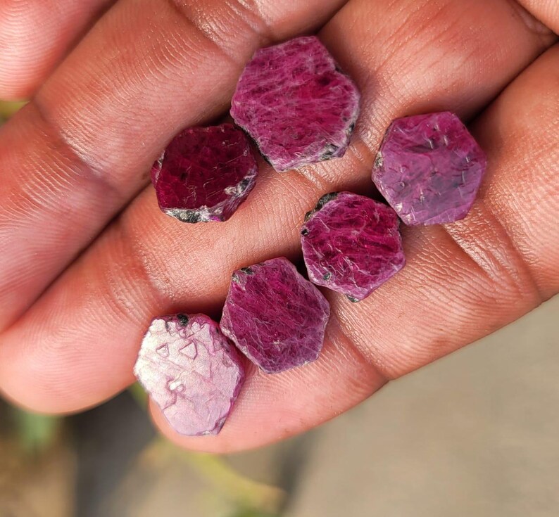 May include: A collection of rough-cut ruby gemstones, each displaying a deep red hue with natural textures. The gemstones vary slightly in size and shape, with some showing hints of lighter pink tones. The gems are held in a hand.