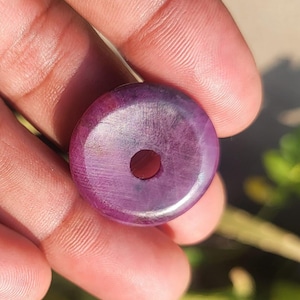 以下が含まれることがあります： 中央に穴の開いた、丸く磨かれた紫色の宝石ビーズ。ビーズは滑らかな表面と深く豊かな色をしています。宝石は手で持たれており、自然光がビーズを照らしています。ビーズはおそらくジュエリー作りに使用されます。