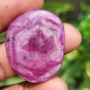 May include: A polished, oval-shaped ruby gemstone. The stone is a deep magenta colour with a hexagonal pattern in the centre. The surface has a textured appearance with small black inclusions.