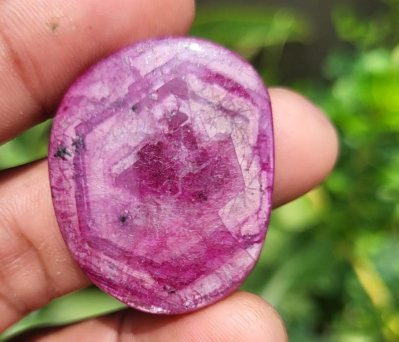 May include: A polished, oval-shaped ruby gemstone. The stone is a deep magenta colour with a hexagonal pattern in the centre. The surface has a textured appearance with small black inclusions.