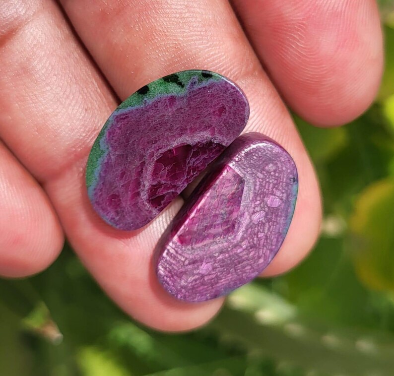 K&ouml;nnte beinhalten: Zwei polierte Edelsteine ​​mit tiefvioletter Farbe, die ein geschichtetes Muster und einen gr&uuml;n-blauen Rand aufweisen. Die Steine ​​werden in einer Hand gehalten, wodurch ihre einzigartige Farbe und Textur zur Geltung kommen. Die Edelsteine ​​sind oval geformt.