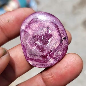 May include: A polished, oval gemstone in a deep magenta colour. The stone is translucent with internal veining and crystalline structures, creating a unique pattern. The surface reflects light, showcasing its faceted design.