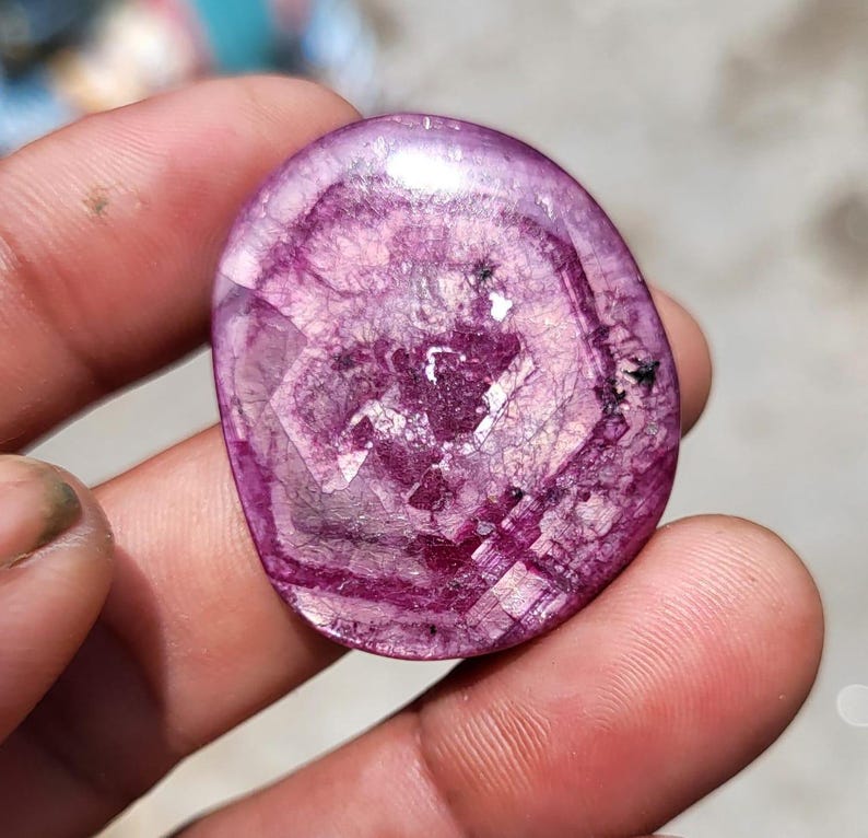 May include: A polished, oval gemstone in a deep magenta colour. The stone is translucent with internal veining and crystalline structures, creating a unique pattern. The surface reflects light, showcasing its faceted design.