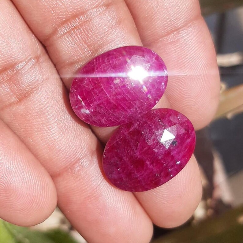 May include: Two oval-shaped, faceted rubies with a deep red hue are displayed. The gemstones are held in a hand, with light reflecting off their surfaces. The rubies have a polished appearance, showcasing their natural beauty.