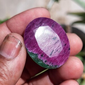 Pode incluir: Gema oval com superfície facetada, exibindo uma vibrante mistura de cores. A gema apresenta um tom magenta profundo com estrias verdes e inclusões pretas. A superfície polida reflete a luz, destacando sua intrincada estrutura interna.