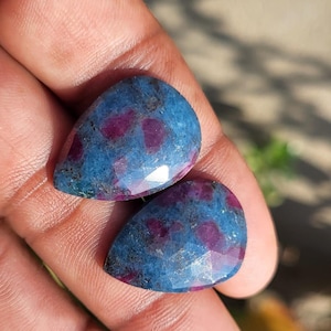Può includere: Due gemme a forma di pera con una superficie sfaccettata. Le pietre hanno un colore di base blu scuro con macchie irregolari di rosso vibrante. Le gemme sono esposte su uno sfondo sfocato.