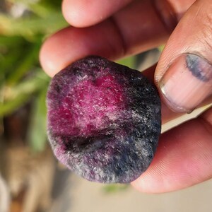 Peut inclure: Gros plan sur une pierre précieuse brute, non taillée, tenue dans une main. La pierre présente un centre magenta profond qui s'estompe en noir et en bords bleu foncé. La surface a un aspect texturé et inégal.