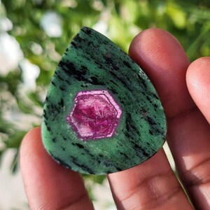 Könnte beinhalten: Ein tropfenförmiger, polierter Edelstein mit einem leuchtend magentafarbenen, hexagonalen Rubinkristall, der in eine gesprenkelte grüne und schwarze Zoisit-Matrix eingebettet ist. Der Stein hat eine glatte, flache Oberfläche mit deutlichem Kontrast.
