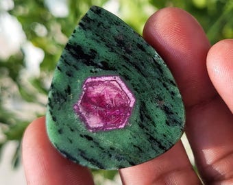 Pode incluir: Uma pedra preciosa polida em forma de lágrima com um cristal de rubi hexagonal magenta vibrante incrustado numa matriz de zoisite verde e preta manchada. A pedra apresenta uma superfície lisa e plana com um contraste de cor distinto.