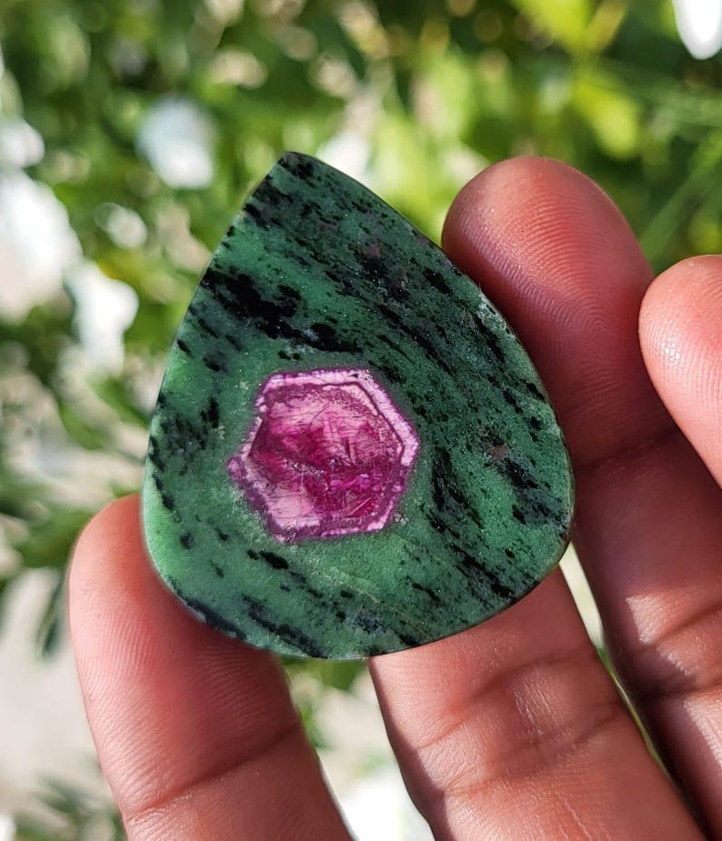 May include: A teardrop-shaped polished gemstone featuring a vibrant magenta hexagonal ruby crystal embedded within a mottled green and black zoisite matrix. The stone displays a smooth, flat surface with distinct colour contrast between the centre.