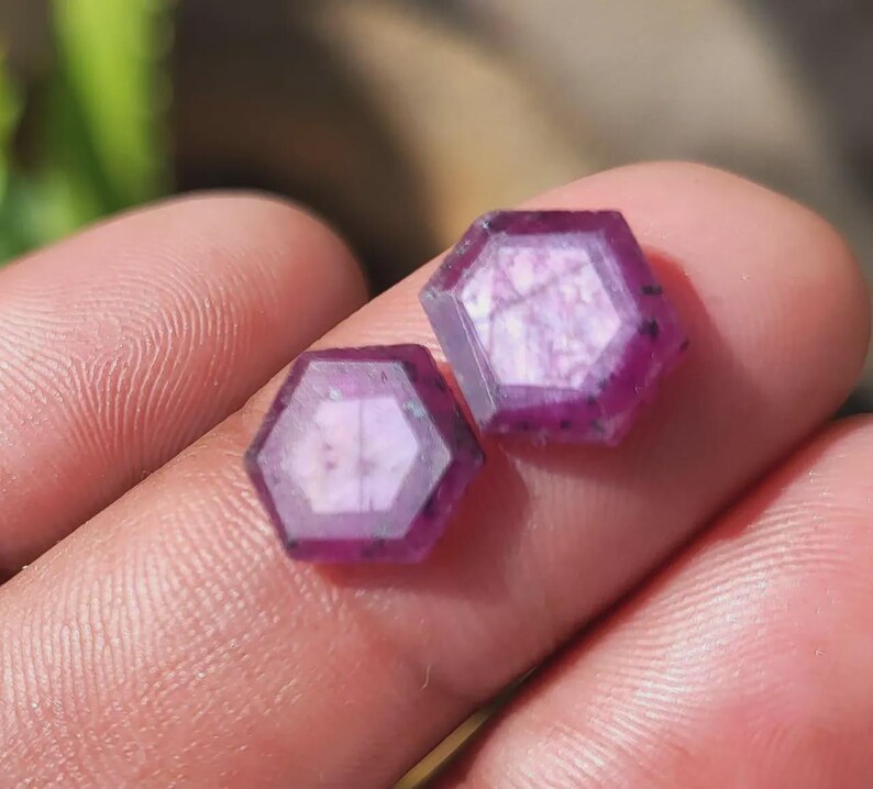 May include: Two hexagonal, faceted gemstones with a deep magenta hue. The gems have a translucent quality, with visible internal patterns and dark inclusions. The edges are sharply defined, and the overall shape is symmetrical.