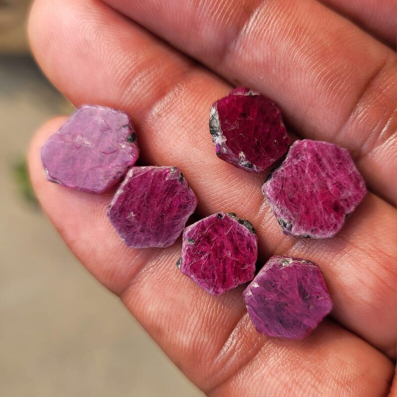 May include: A collection of six raw ruby gemstones. The gemstones are a deep red-pink hue, with visible natural textures and hexagonal shapes. The rubies vary slightly in color and size, showcasing their natural beauty.