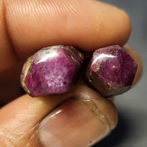 Puede incluir: Dos piedras preciosas de rubí en bruto y sin pulir, con un color rojo magenta intenso y una estructura cristalina hexagonal. Las piedras presentan texturas superficiales naturales, inclusiones visibles y un aspecto semitranslúcido.