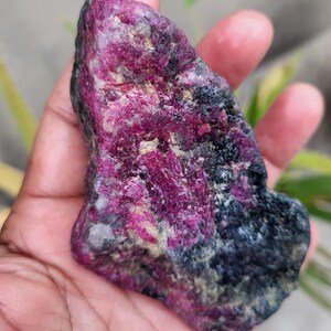Könnte beinhalten: Ein roher, ungeschliffener Edelstein mit einem lebendigen Magenta- und Schwarzfarbschema. Der Stein hat eine strukturierte Oberfläche und wird in einer Hand gehalten. Die Farbe des Edelsteins ist eine Mischung aus tiefen Pink- und Schwarztönen mit helleren Bereichen.