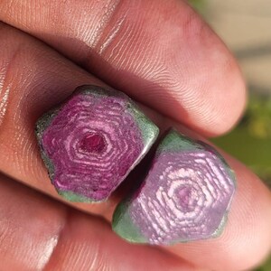 Pode incluir: Duas pedras preciosas em bruto com um padr&atilde;o conc&ecirc;ntrico. As pedras t&ecirc;m um centro magenta profundo, desvanecendo-se para um roxo mais claro com uma borda verde. As pedras preciosas s&atilde;o seguradas em uma m&atilde;o, com um fundo desfocado.