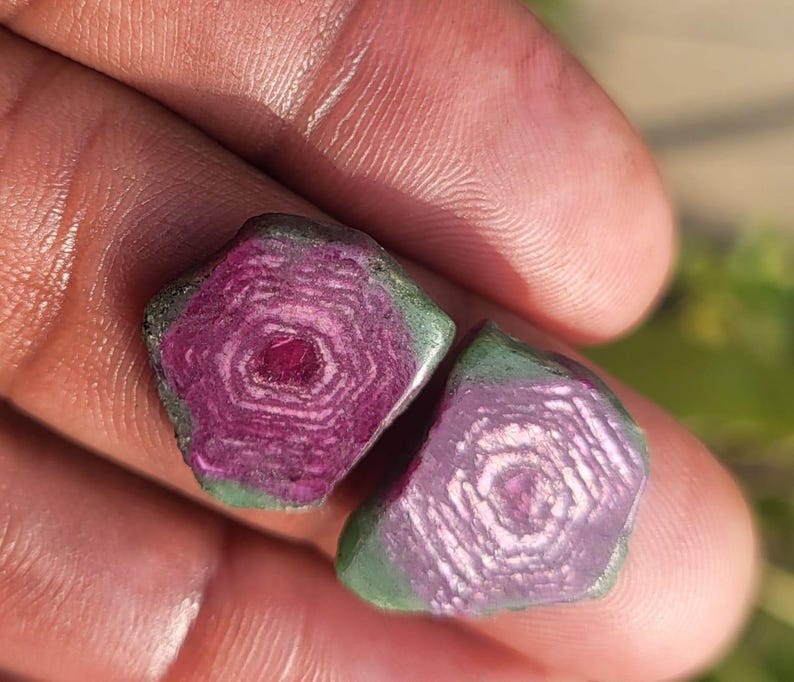 Pode incluir: Duas pedras preciosas em bruto com um padr&atilde;o conc&ecirc;ntrico. As pedras t&ecirc;m um centro magenta profundo, desvanecendo-se para um roxo mais claro com uma borda verde. As pedras preciosas s&atilde;o seguradas em uma m&atilde;o, com um fundo desfocado.