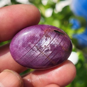 Puede incluir: Una piedra preciosa de rubí pulida de forma ovalada con un tono magenta púrpura intenso. La superficie muestra una textura fibrosa natural distintiva con inclusiones lineales que se cruzan y un acabado suave y reflectante en su cúpula.