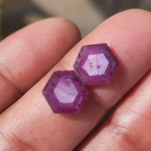 May include: Two faceted, hexagonal gemstones with a deep magenta hue. The gems have a polished, reflective surface and are held in a person's hand. The gems are likely rubies, and are a deep, rich color.
