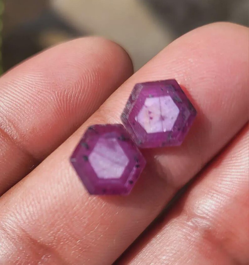 May include: Two faceted, hexagonal gemstones with a deep magenta hue. The gems have a polished, reflective surface and are held in a person's hand. The gems are likely rubies, and are a deep, rich color.